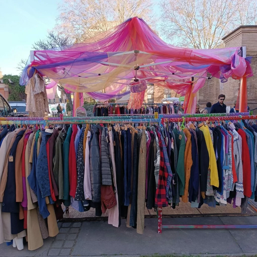Stand de friperie Hyperpop. Tente aux voiles roses, jaunes et violets, portants de vêtements colorés.