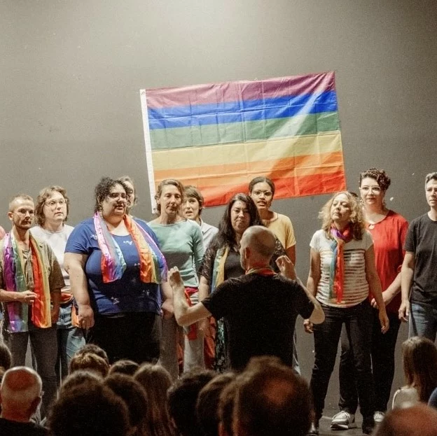 Chorale de l'association Toulouse en Chœurs, Queer à Chœur & Voix des Fiertés, performant devant un drapeau arc-en-ciel