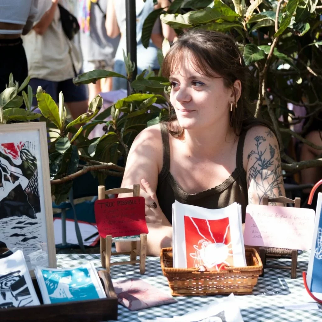 Mélodie Lucas assise à un stand à l'extérieur, exposant ses illustrations et créations.